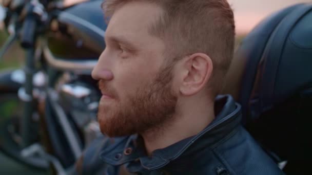 Middle-aged biker with beard sits by motorcycle at sunset — Stock Video