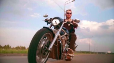 Dövmeli şık bir motorcu gün batımında bir kır yolunda motosiklet sürüyor.