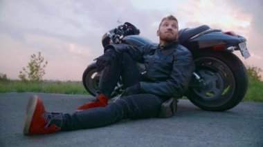 Sakallı orta yaşlı motorcu gün batımında motosikletin yanında oturur.