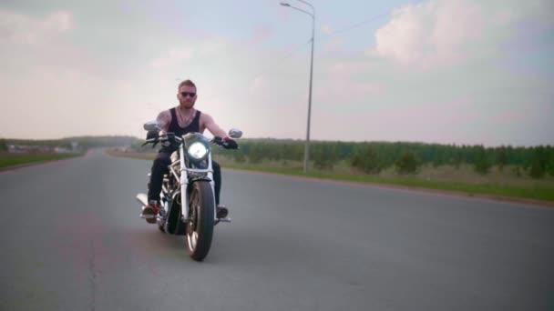 Стильный байкер с татуировками ездит на мотоцикле по проселочной дороге на закате — стоковое видео