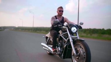 Dövmeli şık bir motorcu gün batımında bir kır yolunda motosiklet sürüyor.