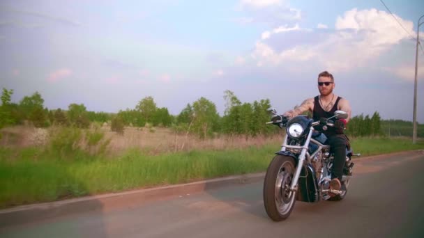 Elegante motociclista com tatuagens monta uma motocicleta em uma estrada rural ao pôr do sol — Vídeo de Stock
