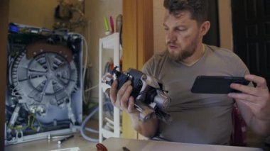 Bir adam çamaşır makinesini tamir eder ve talimatları telefonundan izler.