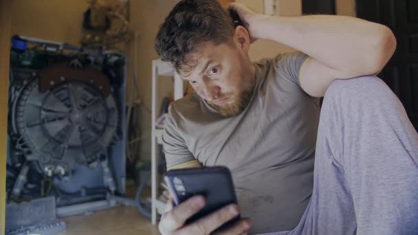 Человек чинит стиральную машину и смотрит инструкции на телефоне — стоковое видео