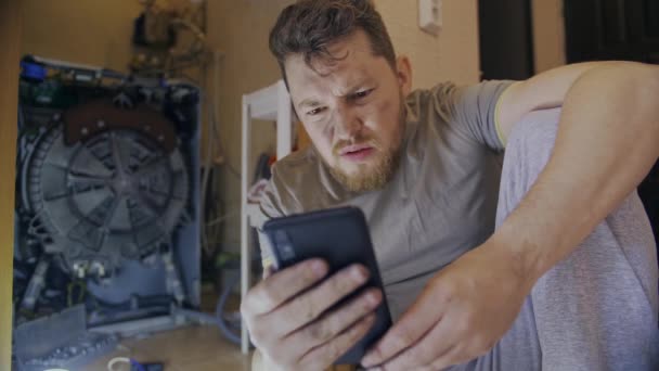 A man fixes a washing machine and watches instructions on his phone — Stock Video