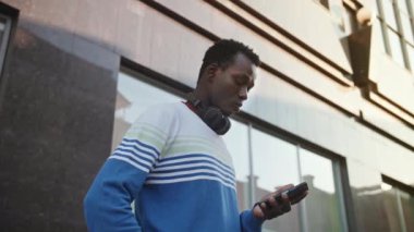 Şık giyinmiş genç siyahi bir öğrenci şehirde oturmuş akıllı bir telefonla konuşuyor.