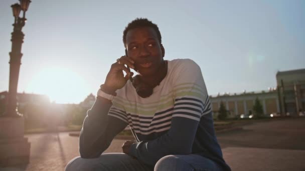 Молодий чорний студент в стильному одязі сидить і розмовляє на смартфоні в місті — стокове відео