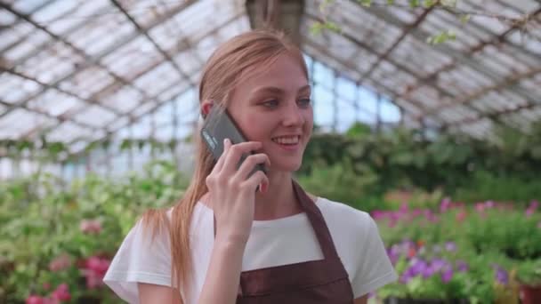 Ung flicka plantskola arbetare och blomma växthus talar med en kund på telefon — Stockvideo