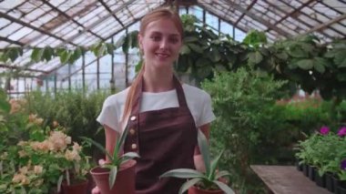 Genç sarışın kız elinde çiçeklerle bir serada yürüyor.