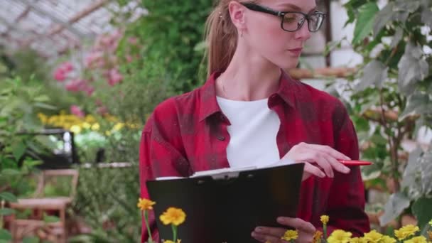 Agronomist meisje in bril en geruit rood shirt controleert de kwaliteit en kwantiteit van de planten in de kas — Stockvideo