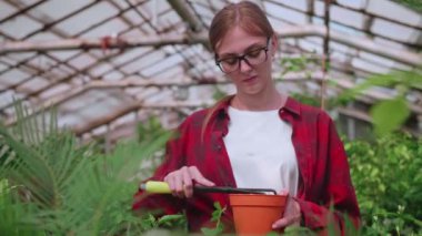 Kırmızı gömlekli kız serada çiçek naklediyor, aletlerle çalışıyor.