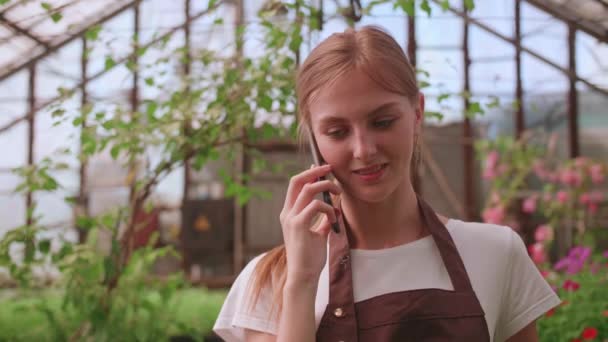 Gadis muda perawat dan bunga rumah kaca berbicara kepada klien di telepon — Stok Video