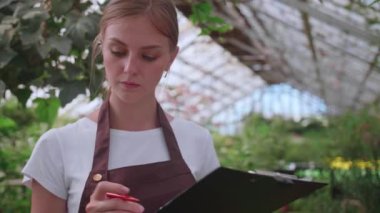 Önlüklü kız müfettiş seradaki çiçekleri kontrol edip sayıyor, kayıtlarını tutuyor.