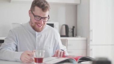 Sabahları gözlüklü genç bir adam kahvaltıda dergi okuyor ve çay içiyor.