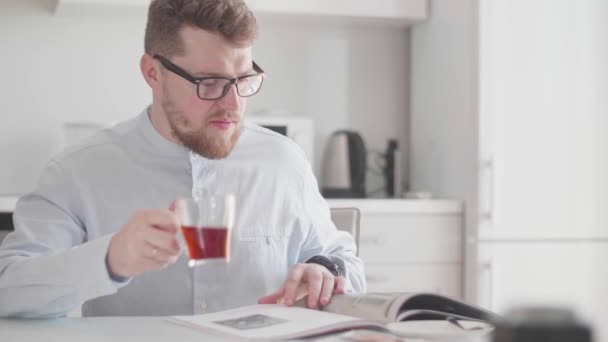 Молодой стильный мужчина в очках утром за завтраком читает журнал и пьет чай — стоковое видео