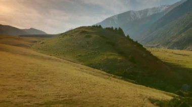 Yeşil tepeler ve tarlalar üzerinde gün batımı ışınları altında uçarak, buzullarla kaplı büyük dağların arka planına karşı. Hafif bir sisi, nehri ve tepesi olan görkemli, huzurlu bir manzara.. 