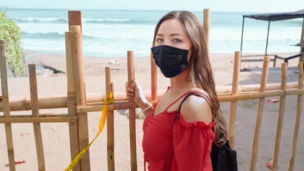 Una hermosa chica con una máscara está de pie cerca de la cerca cerrando la entrada a la playa. Bloqueo.. — Vídeos de Stock