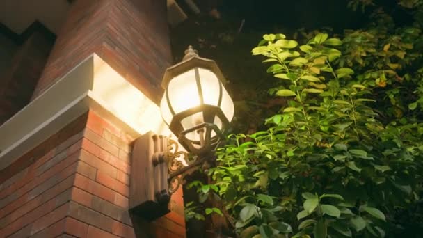 Una hermosa lámpara de calle vintage ubicada en la pared de una antigua casa de ladrillo rojo. La linterna ilumina la calle por la noche con un color amarillo cálido, hay exuberantes arbustos verdes cerca. — Vídeo de stock