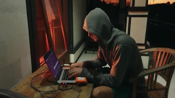 Downshifter Hacker programmerare i en huva skapar ett nytt program med virus. En nybörjare programmerare student arbetar på semester att skapa ny programvara mot bakgrund av en mörk orange solnedgång. — Stockvideo