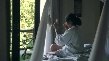 Beyaz cüppeli genç bir kız lüks bir otel odasında geniş pencereli bir yatakta oturuyor. Kız huzurlu bir atmosferde sabah makyajı yapıyor. Ev ücretleri, sabah ücretleri..