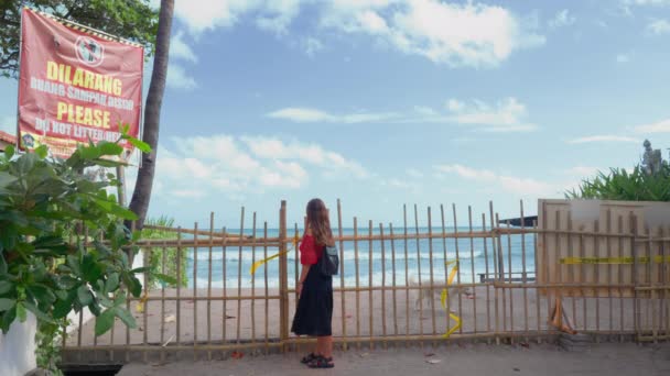 Una niña pequeña en una blusa roja se levanta sobre el fondo de una pared de bambú bloqueando su camino a la playa paradisíaca. — Vídeo de stock