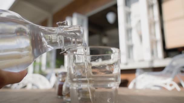 O garçom derrama água de uma garrafa em um copo de vidro transparente em pé sobre a mesa. O terraço de verão do restaurante em um dia ensolarado, sem pessoas. Um restaurante vazio com um cliente. Devagar.. — Vídeo de Stock