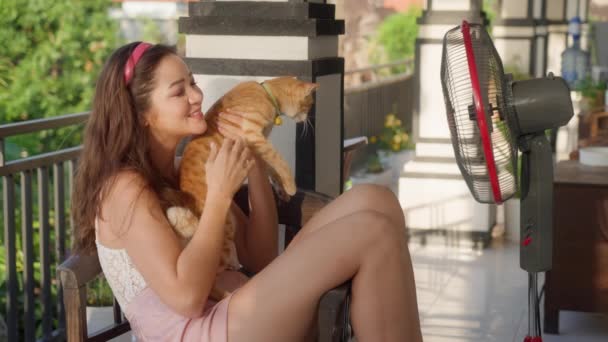 Une jeune fille métissée est assise sur la terrasse avec un chat à côté d'un ventilateur par une chaude journée ensoleillée. L'hôtesse et son mignon chat rouge profitent de l'air frais d'un ventilateur électrique. Réchauffement climatique et étés chauds — Video