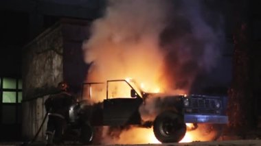 Gizli bir yüzü olan bilinmeyen bir itfaiyeci gece yangın hortumundan çıkan suyla yanan siyah bir jipi söndürüyor. Gece yanan bir araba şehrin arka planında ve sanayi bölgesinde