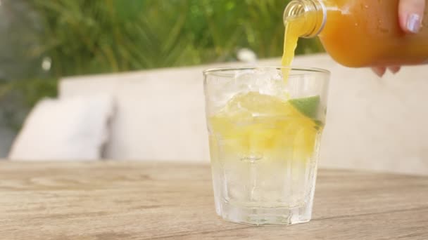 A girl with a beautiful manicure on a sunny day pours orange jamu a natural antibiotic into a glass glass with ice. A close-up of a glass standing on a table in which juice is poured. Slow motion. — Stock Video