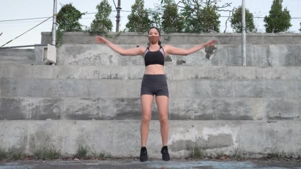 A beautiful girl in a tank top and a medical mask on her face trains during a pandemic on a deserted concrete stadium on a cloudy day. Strong motivation of a girl to play sports in any conditions — Stock Video