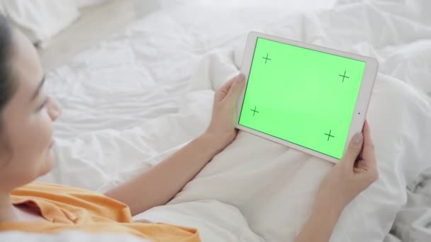 Een meisje met een mooie manicure ligt in een sneeuwwit bed en kijkt naar het scherm van een tablet met een groen scherm en barst in lachen uit. Zelfisolatie. Chromakey scherm voor computer graphics. — Stockvideo