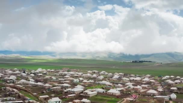 Un paisaje aéreo épico de una pequeña ciudad asentamiento con edificios blancos y un valle verde, bajo enormes nubes de tormenta en un día soleado. Fenómenos naturales únicos. El cuadro fue tomado de un helicóptero en movimiento — Vídeos de Stock