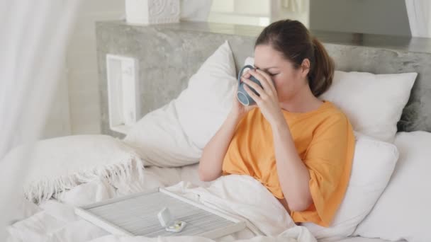 Una chica enferma frustrada de raza mixta está llorando sentada en una cama blanca y bebiendo de una taza azul, una bandeja con medicinas y pastillas está en su regazo. Una chica asiática está enferma en un gran hogar luminoso. — Vídeo de stock