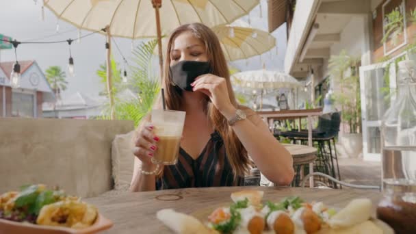 Menina asiática em uma máscara beber coquetel de café tomando palha sob o pano de proteção com piscar lâmpadas elétricas brilhantes no fundo. Coronavirus pandemia tempo vida de luxo em trópicos. — Vídeo de Stock