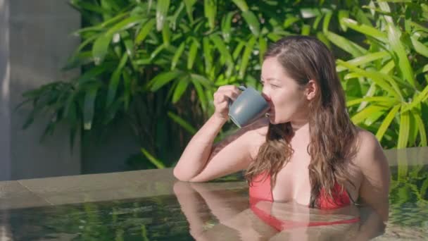 Une belle fille métissée avec de gros beaux seins et un maillot de bain rouge, assise dans la piscine sous les rayons du soleil et buvant dans une tasse bleue faite maison. Une fille asiatique se repose dans une villa. — Video