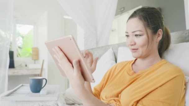 A beautiful mixed-race girl is surprised by a call on a tablet and starts talking via video on a tablet. A successful Woman who is on self-isolation communicates with colleagues sitting in bed. — Stock Video