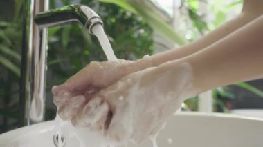 Güzel manikürlü bir kız ellerini lüks bir villada sabunla yıkıyor. Beyaz, modern bir iç mekanda, tropikal yeşil bitkilerin arka planına yakın çekim. Yavaş çekim.