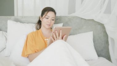 Melez bir tasarımcı kız, parlak, modern bir iç mekanda yatakta yatarken kalemle tablete resim çiziyor. Bir kadın evde salgınla ilgili bir tablet üzerinde çalışıyor..