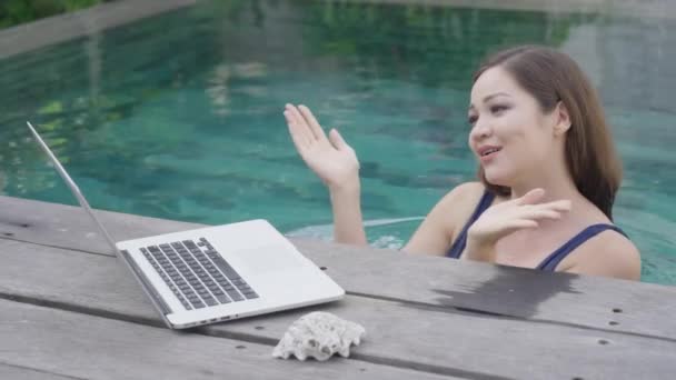 Uma linda garota mestiça está de pé na piscina e conversando via link de vídeo através de um laptop. Uma garota freelance em um maiô azul sorri e mostra um sinal de coração com gestos de mão, envia beijos de ar. — Vídeo de Stock