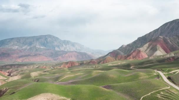 大きな山と雲と青い空を背景に、赤い岩や砂の丘の上に緑の牧草地とユニークな空中風景。岩の間に道路がある絵のような飛行. — ストック動画
