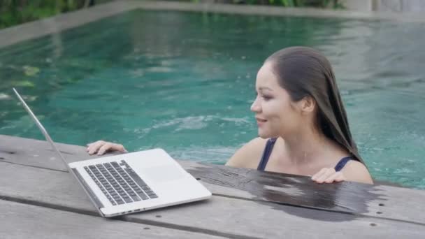 Une belle fille métissée émerge de la piscine et répond à un appel vidéo sur son ordinateur portable. une programmeuse indépendante parle par vidéocommunication avec ses collègues de travail. Mouvement lent. — Video