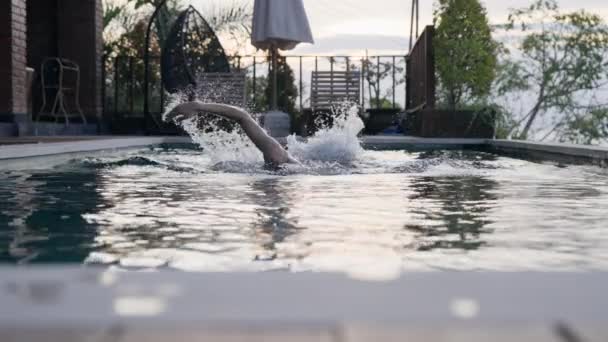 Une personne nage dans la piscine dans une villa chère avec une belle vue sur le ciel couchant. Un homme nage dans une piscine au ralenti, les mains et les éclaboussures d'eau apparaissent hors de l'eau. — Video