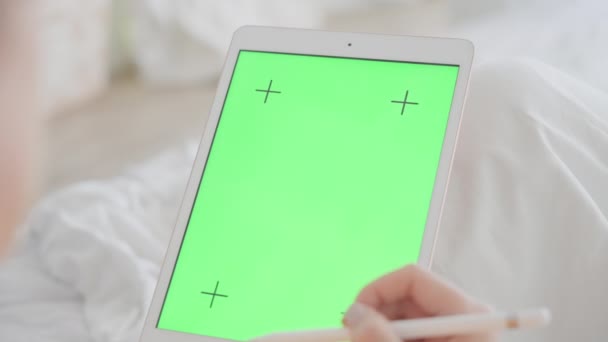 Un tablet con uno schermo verde per la grafica. Una donna con una bella manicure rosa scorre le pagine Internet su un tablet e mette mi piace. Utilizza uno stilo, sullo sfondo di un letto bianco. — Video Stock