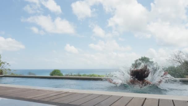 Une belle jeune fille aux cheveux longs et un maillot de bain bleu saute dans la piscine au ralenti, dans une villa chère avec une belle vue sur fond de ciel bleu et de nuages blancs sur une journée. — Video