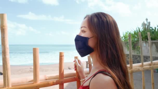 Maskeli güzel bir genç kız bambu çitin önünde duruyor ve karantinaya alınmış sahile bakıyor.. — Stok video