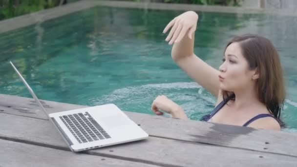 Una hermosa chica mestiza está de pie en la piscina y bailando delante de un portátil. Una chica independiente en traje de baño azul baila después de ver los resultados de inversiones exitosas en una pantalla de computadora — Vídeos de Stock
