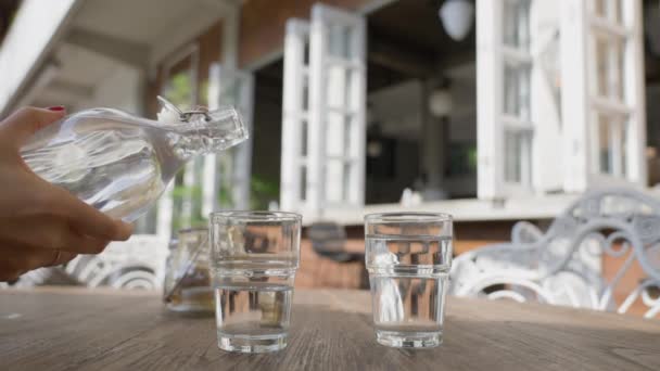 Seorang wanita dengan manikur merah menuangkan air dari botol ke gelas transparan berdiri di atas meja. Teras musim panas restoran pada hari yang cerah, tanpa orang-orang. — Stok Video