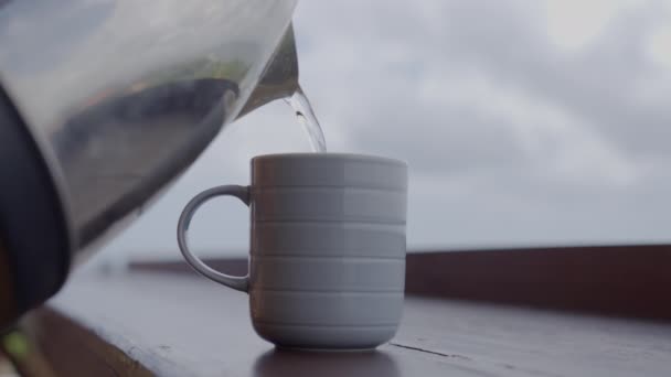 Osoba wylewa gorącą wodę z chromowanego czajnika do niebieskiego kubka na drewnianej podstawce na tle zachmurzonego nieba w ciągu dnia. Zbliżenie zaparzania herbaty strzał w zwolnionym tempie — Wideo stockowe