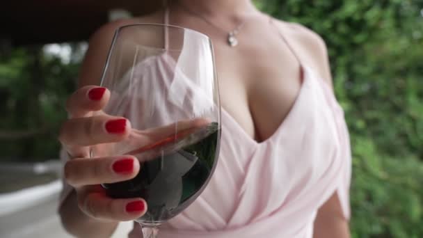 Een heel mooi jong meisje in een zachte roze avondjurk met een grote hals en een rode manicure schudt wijn in een glas Een vrouw met grote borsten zwaait een glas rode wijn close-up in een langzame — Stockvideo