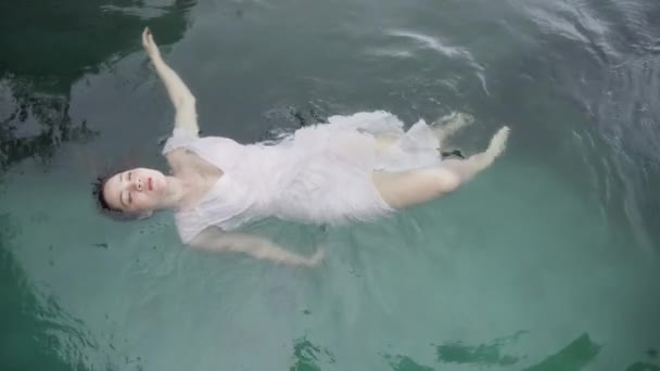 Draufsicht auf ein Mischlingsmädchen in einem zartrosa Abendkleid, das bei trübem Wetter auf ihrem Rücken in einem Pool mit smaragdgrünem Wasser schwimmt. Ein asiatisches Mädchen liegt auf dem Rücken in einem Pool. — Stockvideo